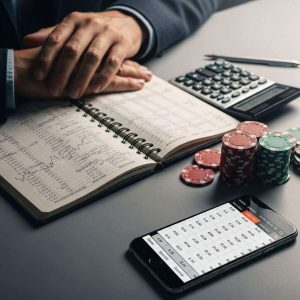 A close-up of a bettor analyzing a notebook with betting records and a calculator, alongside chips and a smartphone displaying betting odds